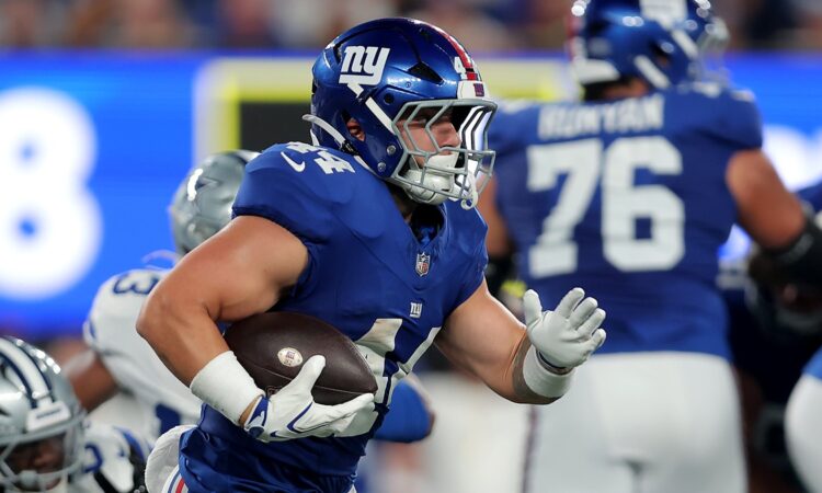 Giants running back Cam Skattebo tucks the ball and cuts upfield with a Cowboys defender trailing behind him.