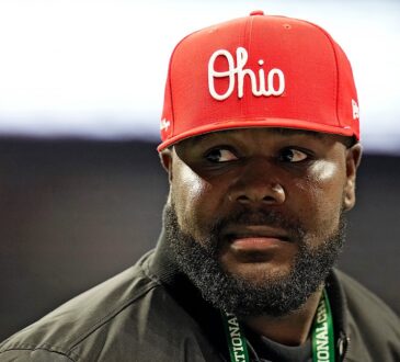 Former Ohio State quarterback Cardale Jones, in a red Ohio cap and a CFP credential lanyard, looks off to his left on the sideline.