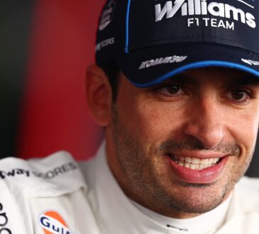 Williams driver Carlos Sainz smiles in a team cap and race suit during a close-up portrait.
