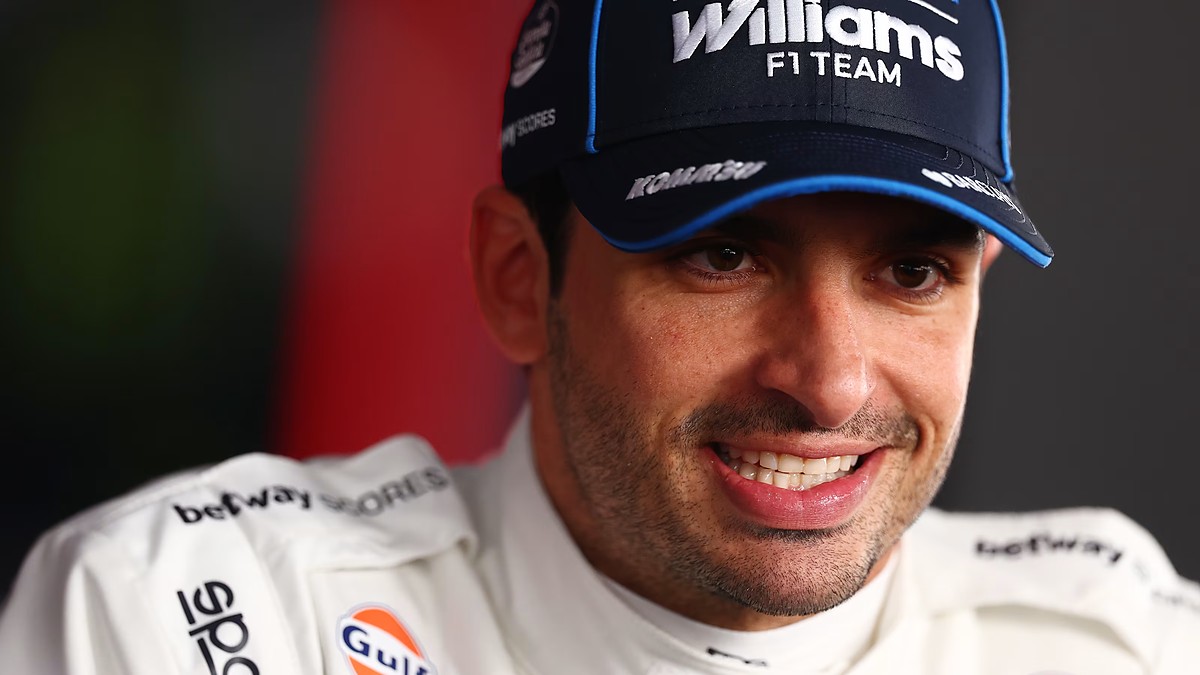 Williams driver Carlos Sainz smiles in a team cap and race suit during a close-up portrait.