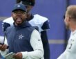 Dallas Cowboys defensive coordinator Christian Parker talks while holding a radio and a call sheet at a joint practice.