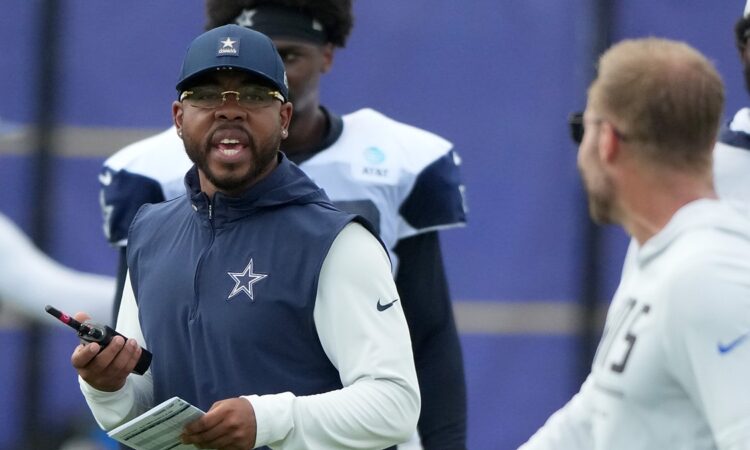 Dallas Cowboys defensive coordinator Christian Parker talks while holding a radio and a call sheet at a joint practice.