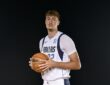 Dallas Mavericks forward Cooper Flagg holds a basketball against a black backdrop in a white Dallas jersey.