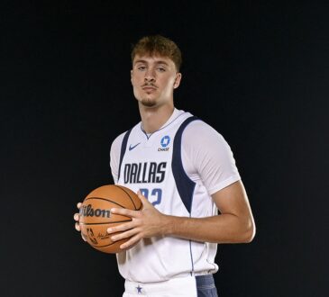 Dallas Mavericks forward Cooper Flagg holds a basketball against a black backdrop in a white Dallas jersey.