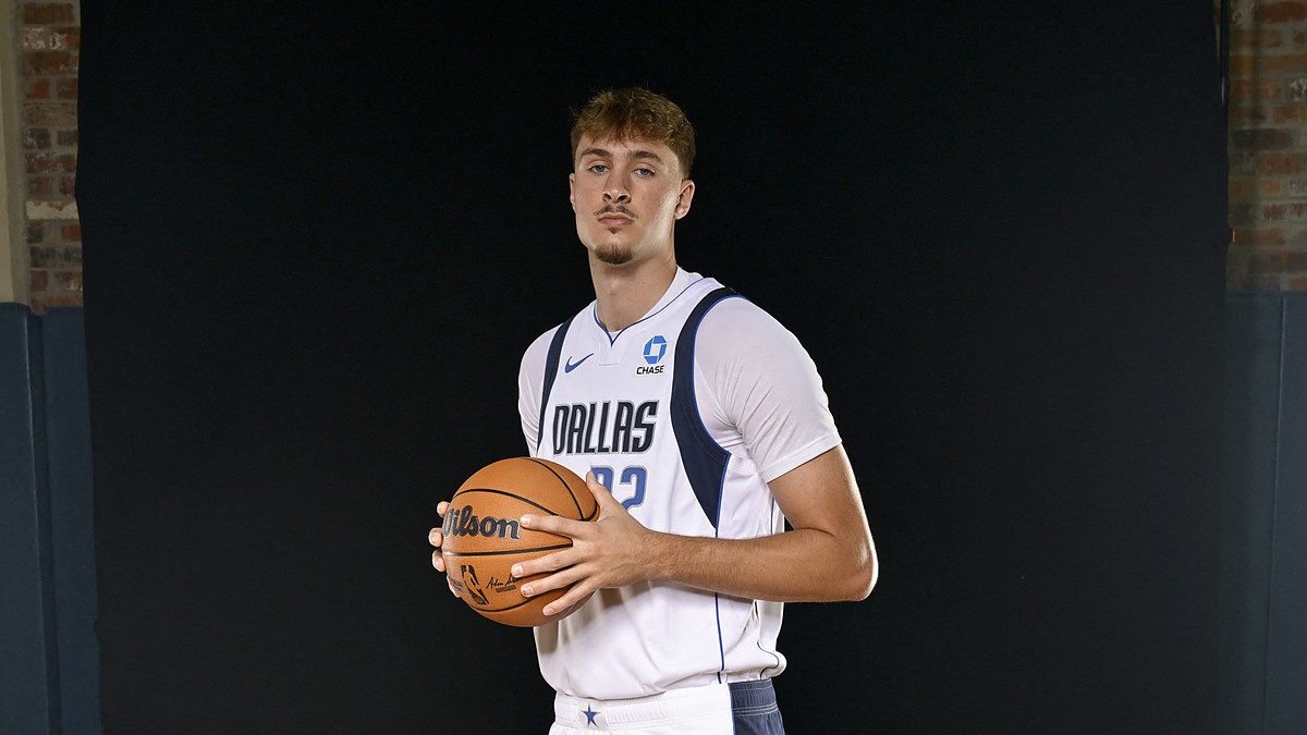 Dallas Mavericks forward Cooper Flagg holds a basketball against a black backdrop in a white Dallas jersey.