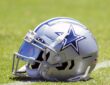 A Dallas Cowboys helmet with a blue mirrored visor sits on the grass at training camp.