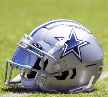 A Dallas Cowboys helmet with a blue mirrored visor sits on the grass at training camp.