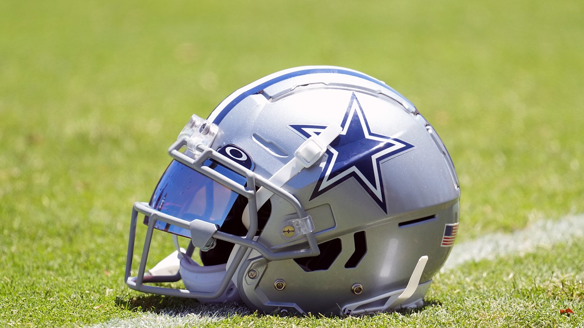 A Dallas Cowboys helmet with a blue mirrored visor sits on the grass at training camp.