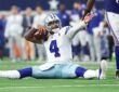 Alt Text: Dallas Cowboys quarterback Dak Prescott sits on the turf holding the ball and raises an arm as a Giants defender walks past.