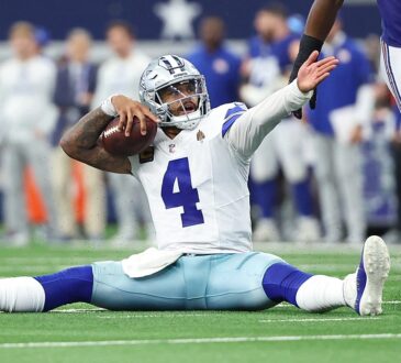 Alt Text: Dallas Cowboys quarterback Dak Prescott sits on the turf holding the ball and raises an arm as a Giants defender walks past.