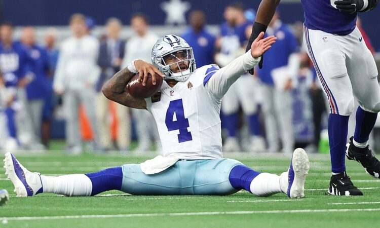 Alt Text: Dallas Cowboys quarterback Dak Prescott sits on the turf holding the ball and raises an arm as a Giants defender walks past.