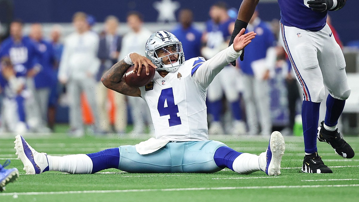 Alt Text: Dallas Cowboys quarterback Dak Prescott sits on the turf holding the ball and raises an arm as a Giants defender walks past.