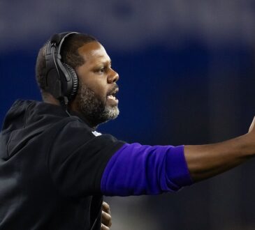Coach Daronte Jones extends an arm and calls out instructions from the sideline in a headset.