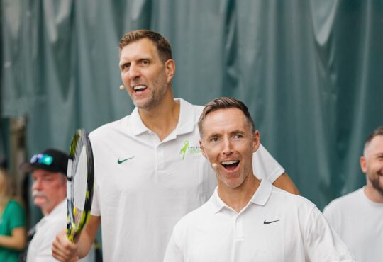 Former Dallas Mavericks forward Dirk Nowitzki smiles and high-fives an opponent across the net at a celebrity tennis match.