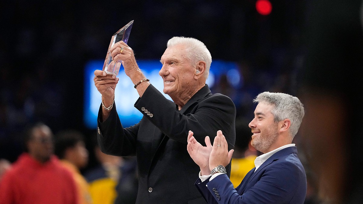 Former Dallas Mavericks coach Don Nelson smiles and raises a crystal trophy as a man beside him applauds.