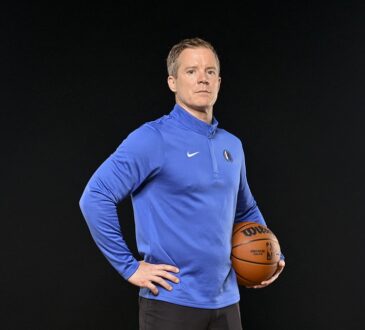 Dallas Mavericks head coach Dusty May stands with a hand on his hip and a basketball under his arm.