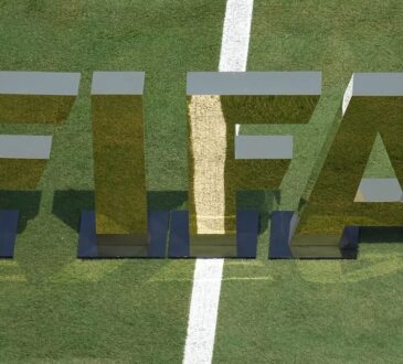 Large reflective FIFA letters standing on a grass pitch beside a painted white line.
