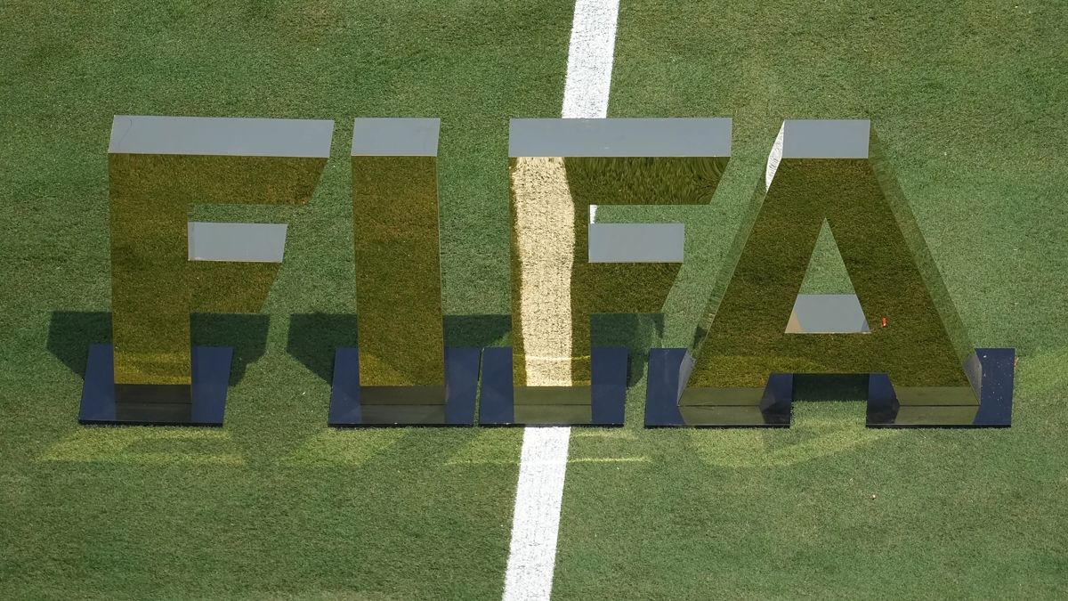 Large reflective FIFA letters standing on a grass pitch beside a painted white line.