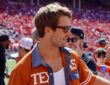 Actor Glen Powell, wearing a burnt orange Texas shirt and sunglasses, smiles while talking with a woman on the field.