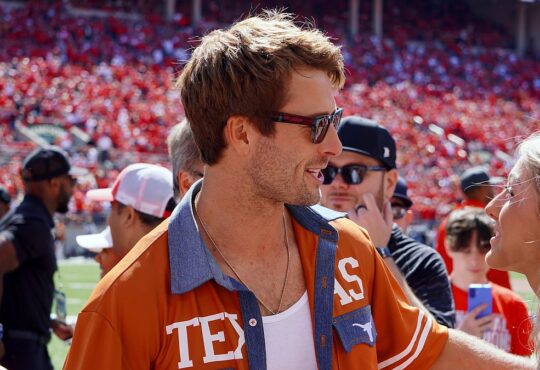 Actor Glen Powell, wearing a burnt orange Texas shirt and sunglasses, smiles while talking with a woman on the field.