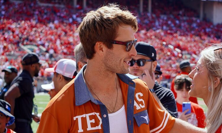 Actor Glen Powell, wearing a burnt orange Texas shirt and sunglasses, smiles while talking with a woman on the field.