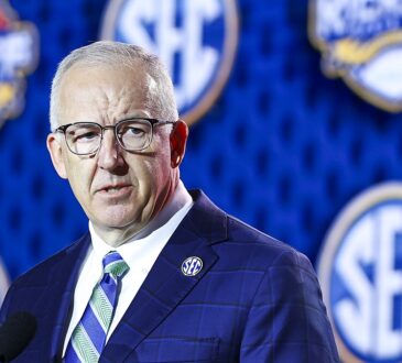 SEC Commissioner Greg Sankey speaks at a podium in front of a wall of conference logos.
