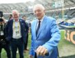 Dallas Cowboys owner Jerry Jones smiles on the field in a light blue sport coat as people around him look on.