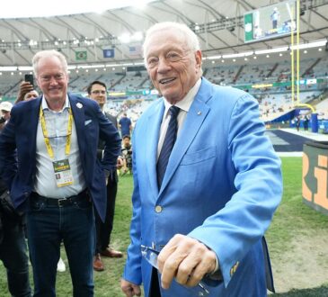 Dallas Cowboys owner Jerry Jones smiles on the field in a light blue sport coat as people around him look on.