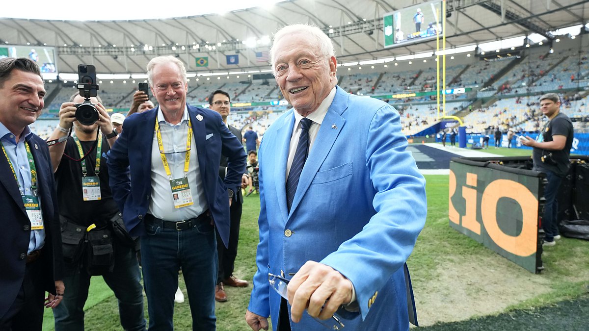 Dallas Cowboys owner Jerry Jones smiles on the field in a light blue sport coat as people around him look on.