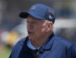 Dallas Cowboys owner Jerry Jones stands on the field in a team cap and jacket at training camp.