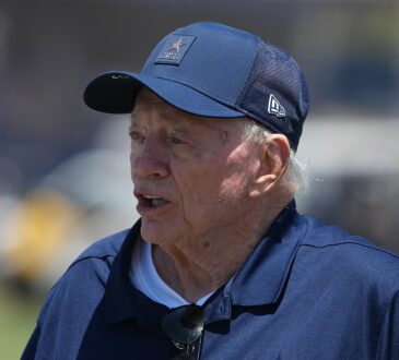 Dallas Cowboys owner Jerry Jones stands on the field in a team cap and jacket at training camp.