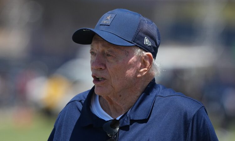Dallas Cowboys owner Jerry Jones stands on the field in a team cap and jacket at training camp.