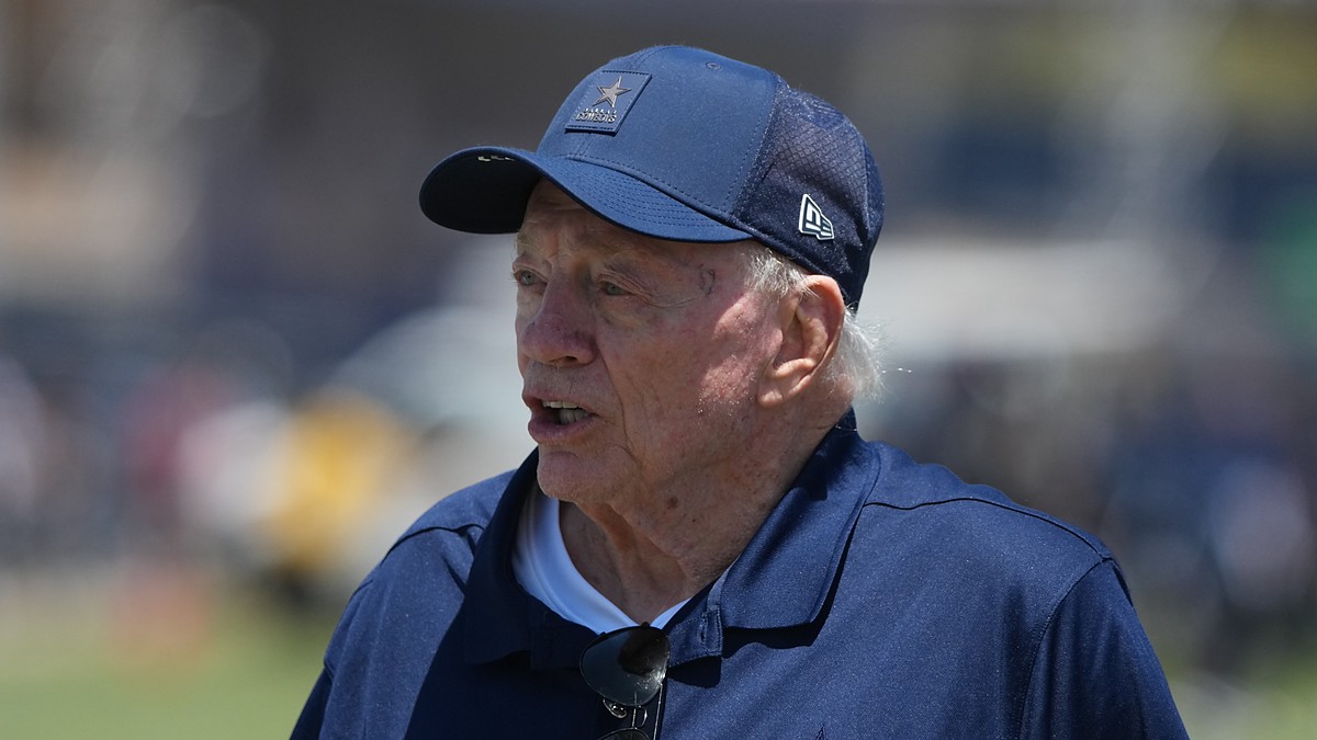 Dallas Cowboys owner Jerry Jones stands on the field in a team cap and jacket at training camp.