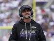 Baltimore Ravens head coach Jesse Minter smiles on the sideline while wearing a headset and holding a folder.