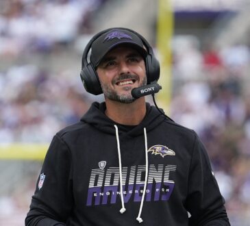 Baltimore Ravens head coach Jesse Minter smiles on the sideline while wearing a headset and holding a folder.