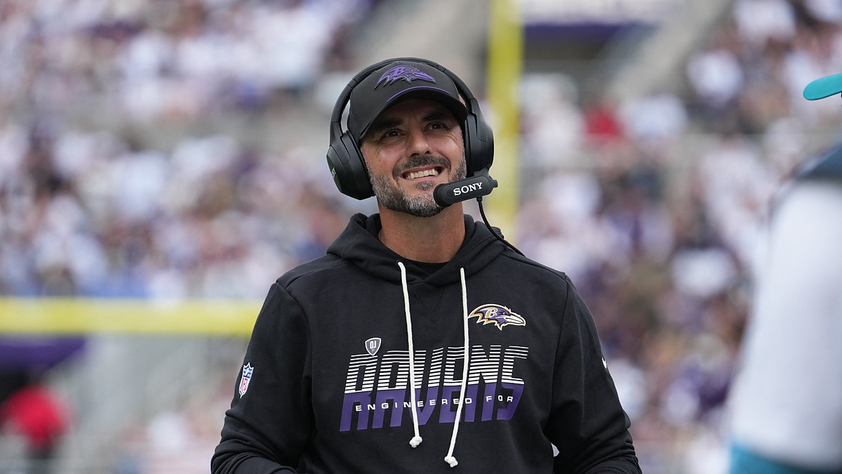 Baltimore Ravens head coach Jesse Minter smiles on the sideline while wearing a headset and holding a folder.