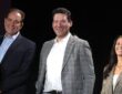 CBS Sports broadcasters Jim Nantz, Tony Romo, and Tracy Wolfson stand together onstage, all smiling.