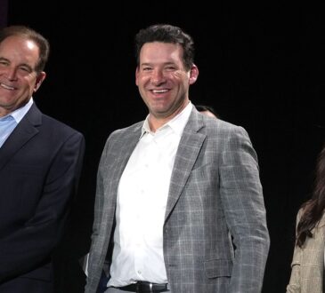 CBS Sports broadcasters Jim Nantz, Tony Romo, and Tracy Wolfson stand together onstage, all smiling.