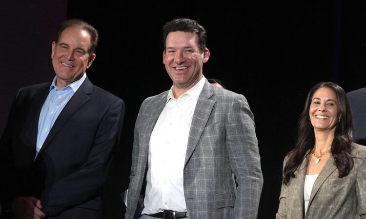 CBS Sports broadcasters Jim Nantz, Tony Romo, and Tracy Wolfson stand together onstage, all smiling.