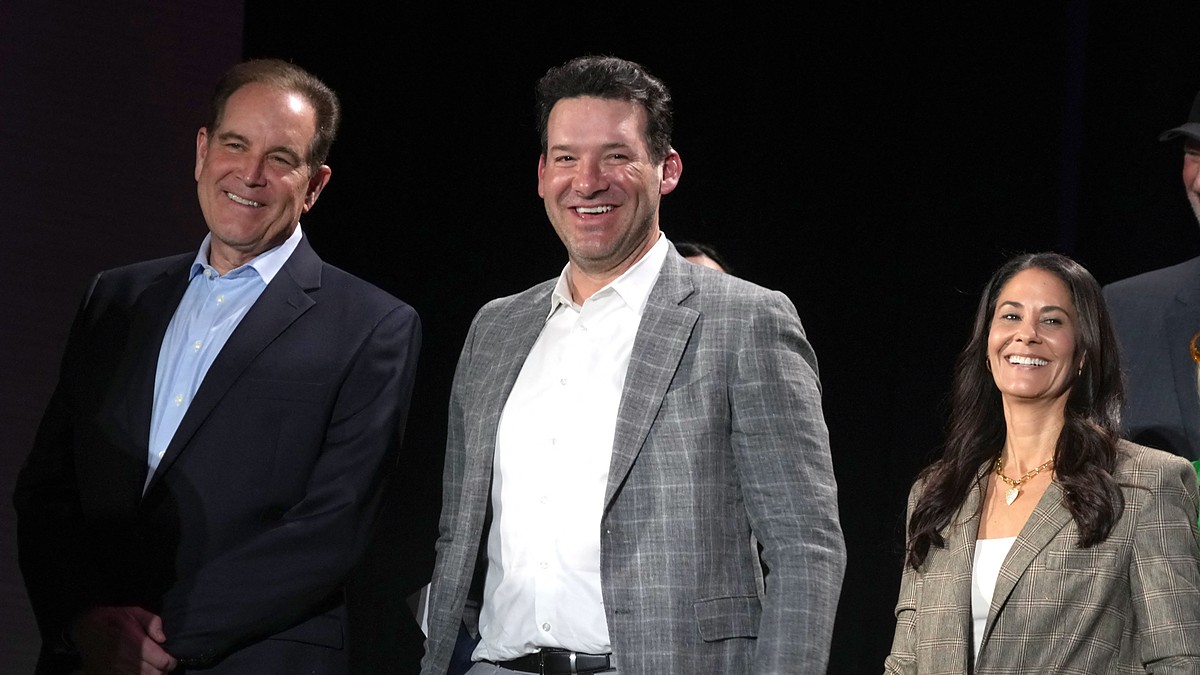 CBS Sports broadcasters Jim Nantz, Tony Romo, and Tracy Wolfson stand together onstage, all smiling.