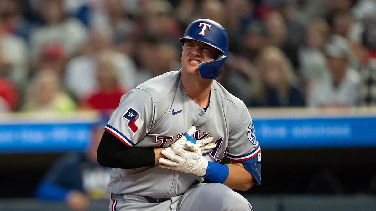 Texas Rangers designated hitter Josh Jung kneels near home plate, grimacing and clutching his gloved hand to his chest.