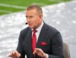 Alt Text: ESPN analyst Kirk Herbstreit gestures with both hands while talking on the broadcast set beside the field.