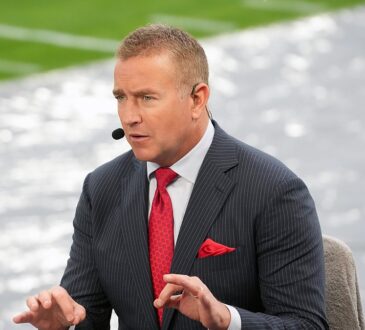 Alt Text: ESPN analyst Kirk Herbstreit gestures with both hands while talking on the broadcast set beside the field.