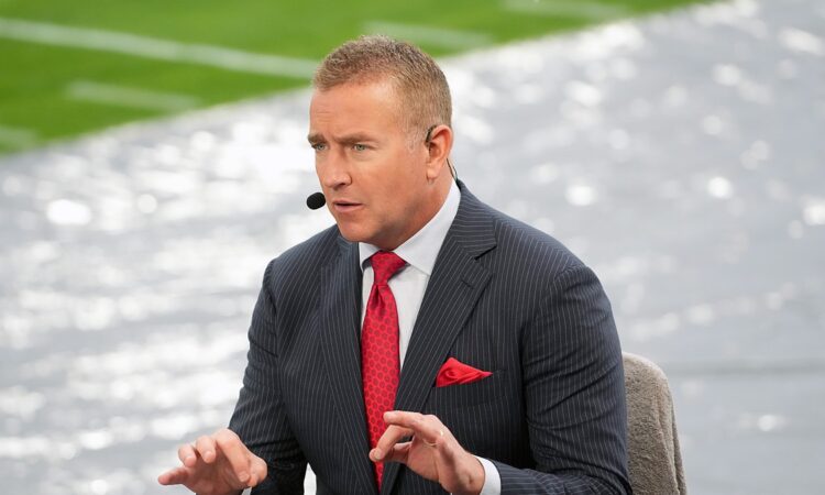 Alt Text: ESPN analyst Kirk Herbstreit gestures with both hands while talking on the broadcast set beside the field.