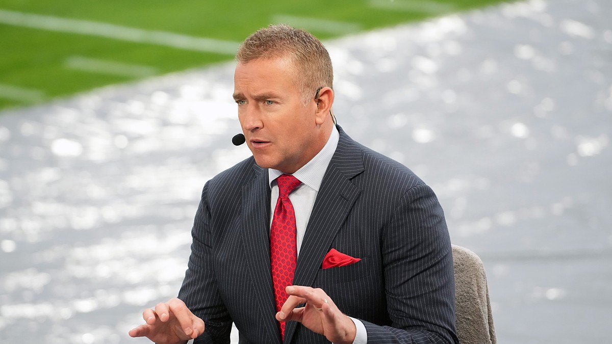 Alt Text: ESPN analyst Kirk Herbstreit gestures with both hands while talking on the broadcast set beside the field.