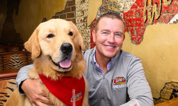 Kirk Herbstreit sits in a booth with his golden retriever Peter, who wears a red Raising Cane's bandana.
