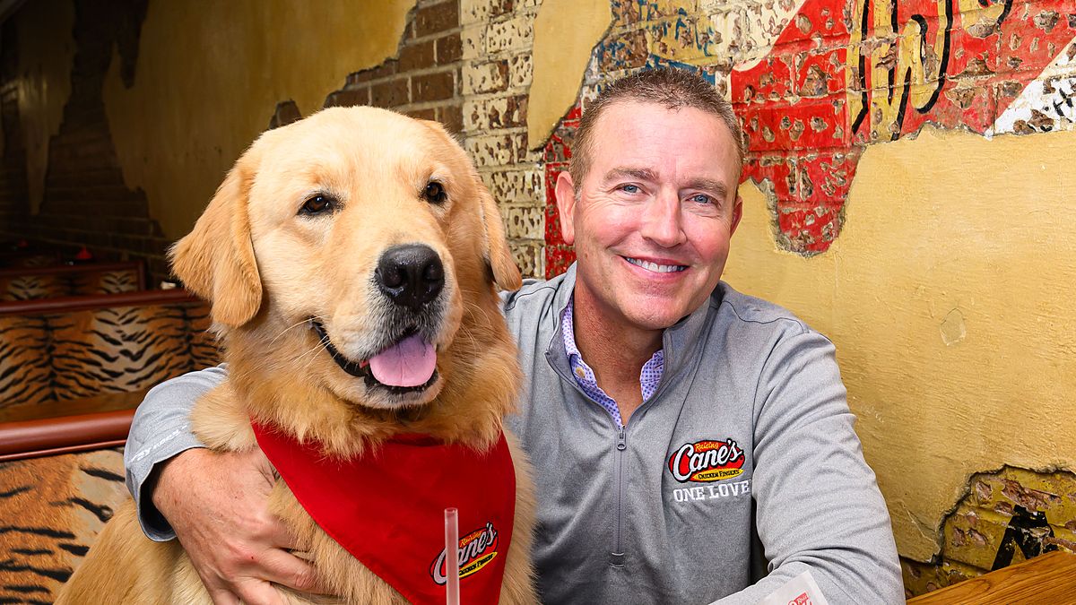 Kirk Herbstreit sits in a booth with his golden retriever Peter, who wears a red Raising Cane's bandana.