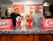 Todd Graves and Kirk Herbstreit stand behind a Raising Cane's counter with two dogs in Cane's bandanas.