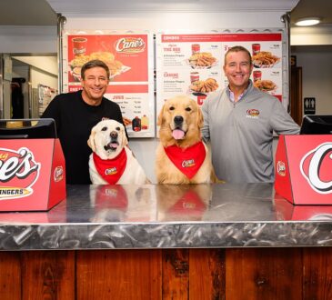 Todd Graves and Kirk Herbstreit stand behind a Raising Cane's counter with two dogs in Cane's bandanas.