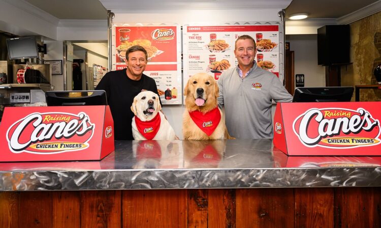 Todd Graves and Kirk Herbstreit stand behind a Raising Cane's counter with two dogs in Cane's bandanas.